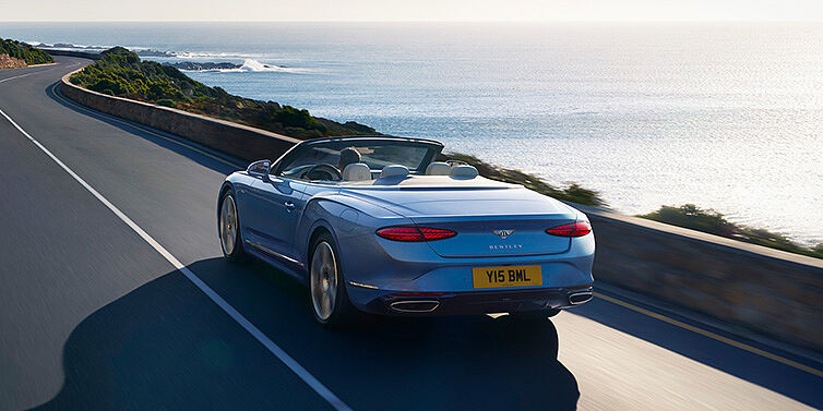 Bentley Madrid New Bentley Continental GTC Azure convertible rear view in Light Windsor blue paint with 22 inch ten spoke wheels driving dynamically on an ocean road in the sunshine